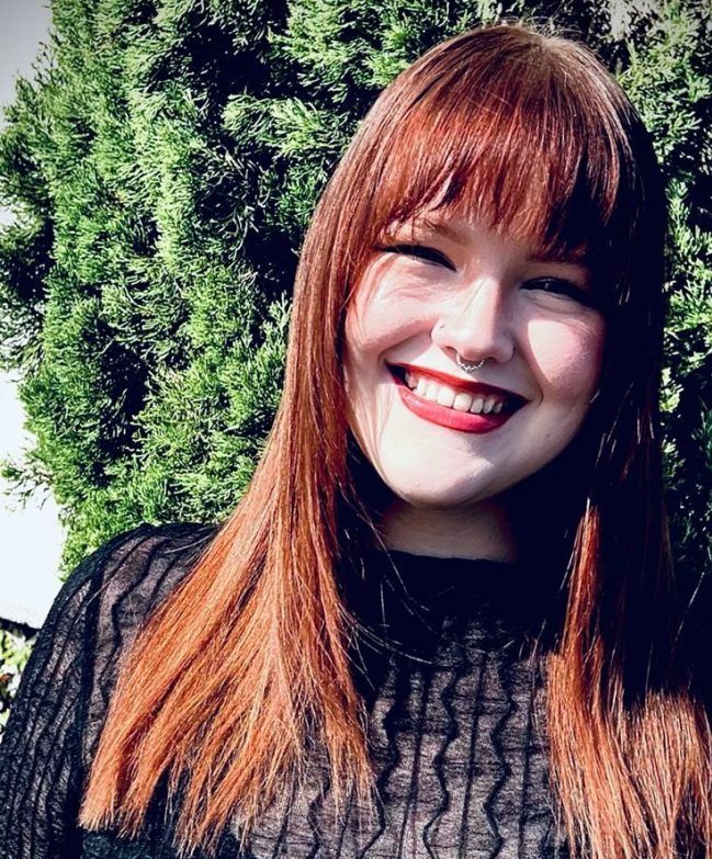 Woman with red hair and bangs smiling outdoors, wearing a black top, and a nose ring.