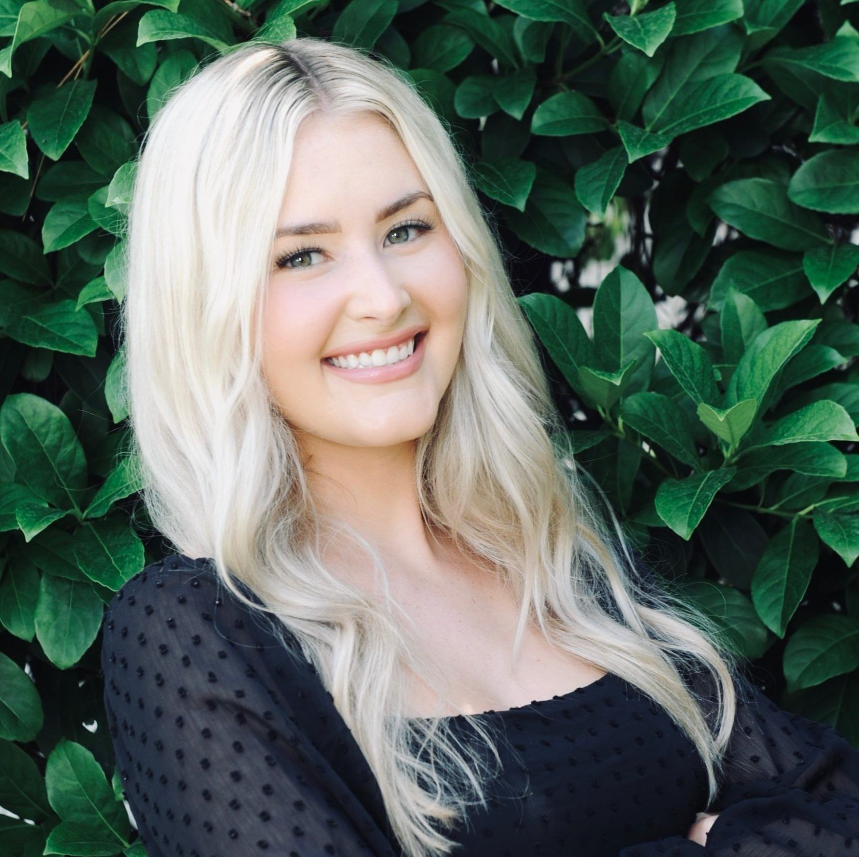 Blonde woman with a wide smile, wearing a black top, in front of green foliage.