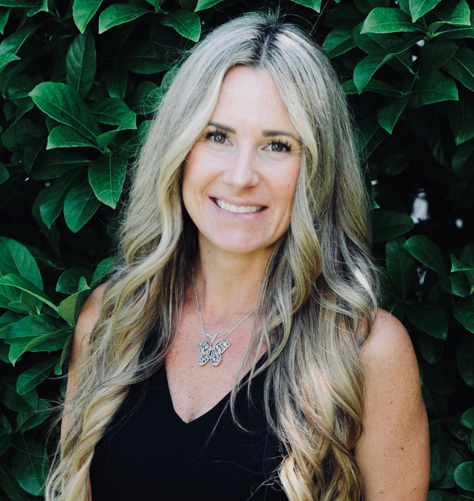 Woman with long blonde hair smiles, wearing a black top and silver necklace, against a green leafy background.