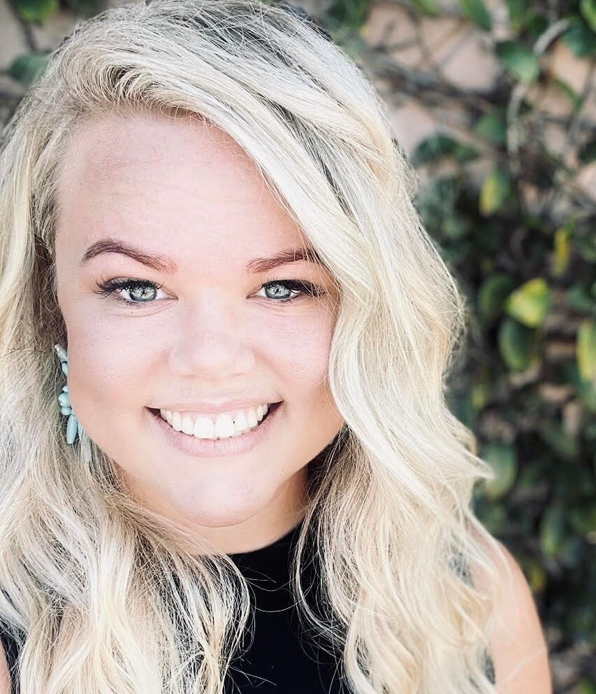 Blonde woman smiles at the camera. She has light eyes and a black top. Background: green leaves.