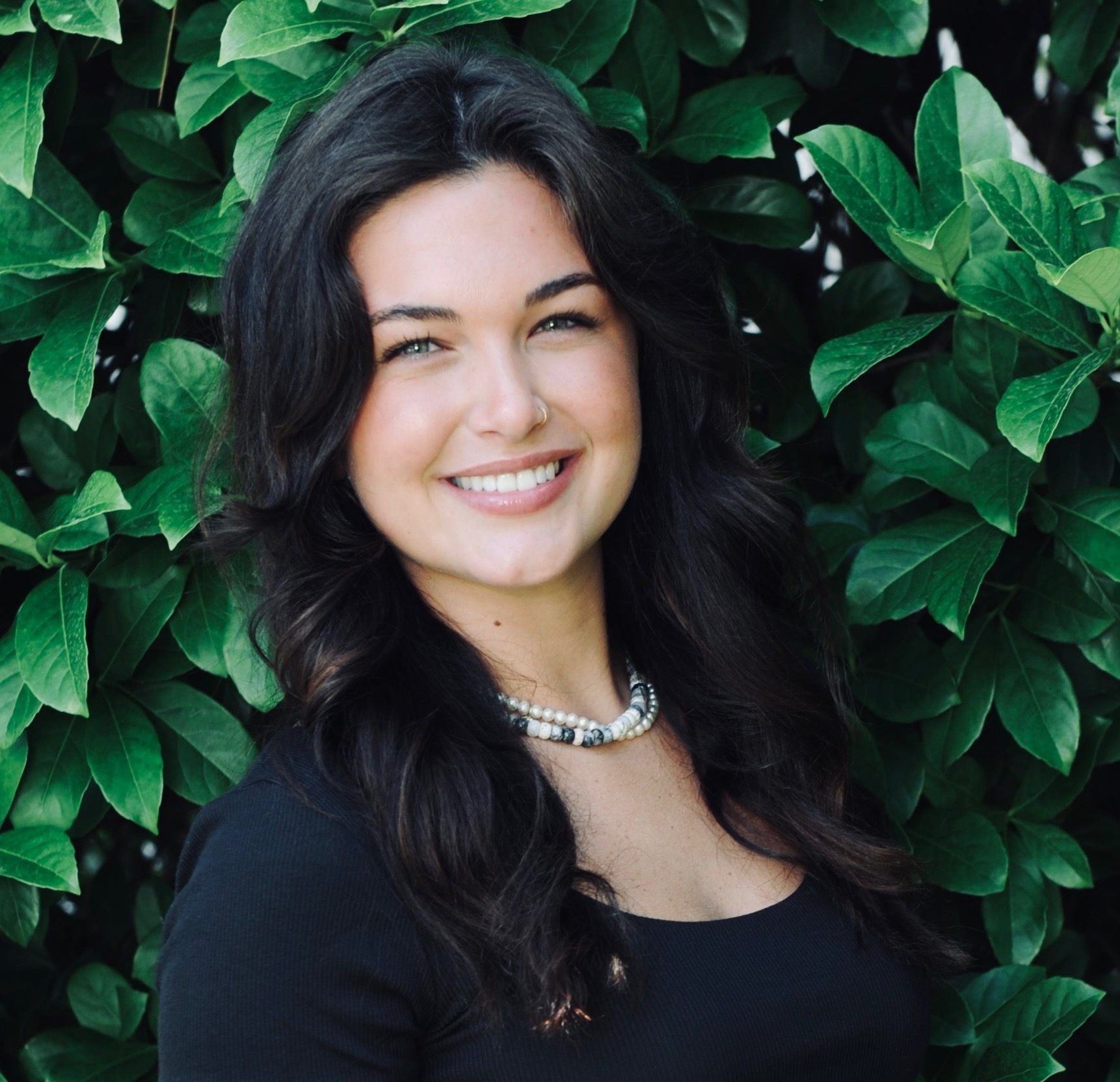 Woman with dark hair smiles at the camera. She's wearing a dark top and pearl necklace, in front of green leaves.