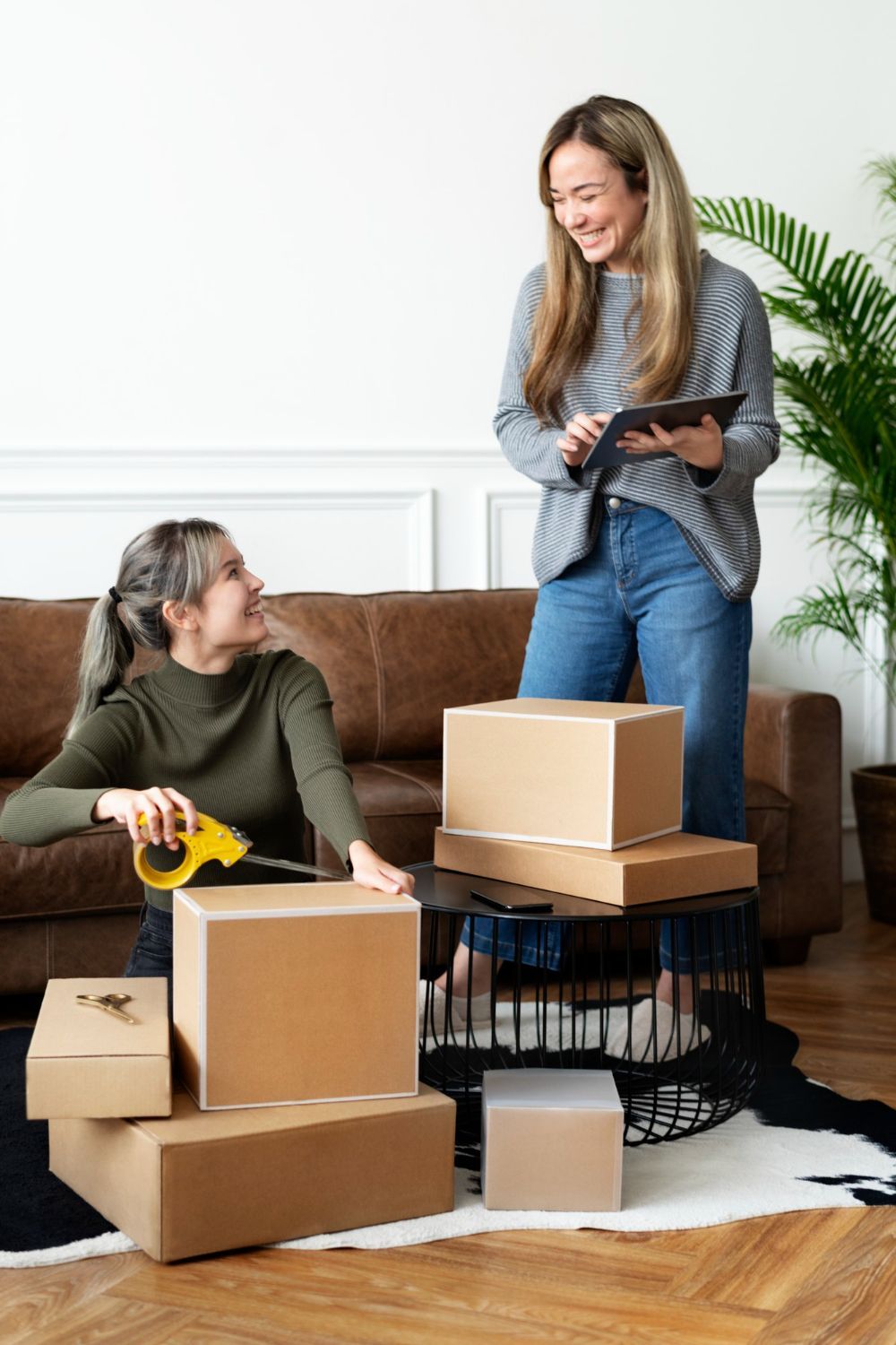 Two women are packing boxes in a living room.