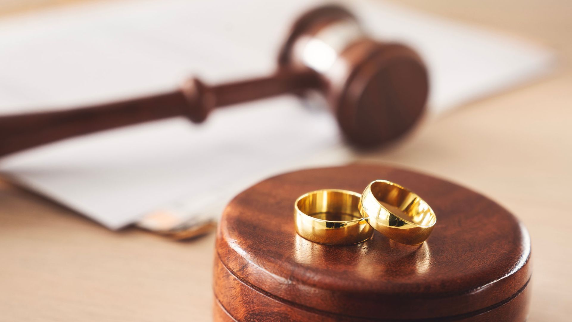 Two gold wedding bands rest on a wooden stand, with a gavel and paperwork blurred in the background.