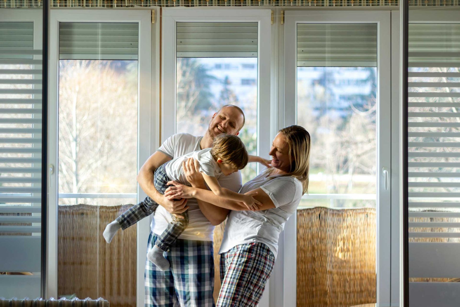 Family smiling and embracing in front of window. Child in mid-air, playing, wearing pajamas. Family smiling and embracing in front of window. Child in mid-air, playing, wearing pajamas.
