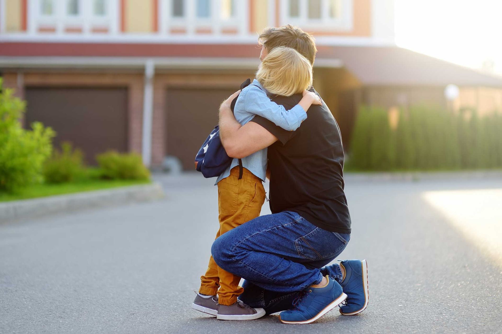 Father hugging a child, kneeling on a driveway in front of a house.