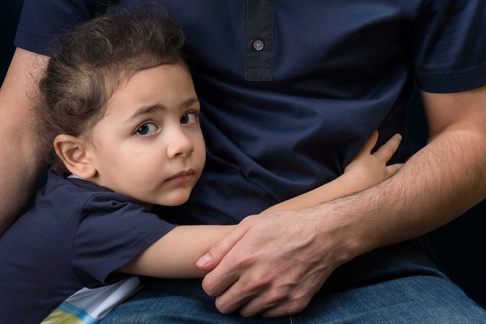 Child hugging a person wearing a navy blue shirt and jeans. The child has a worried expression.