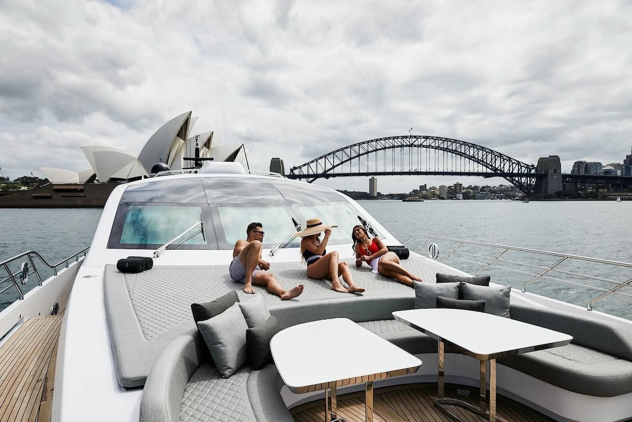 Three people lounge on the deck of a yacht in Sydney Harbour, with the Opera House and Harbour Bridge in the background.