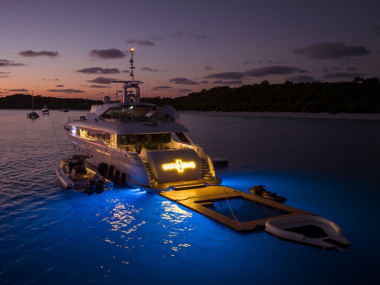 A luxury yacht at dusk, illuminated with bright blue underwater lights, docked near a beach with an attached swim platform.