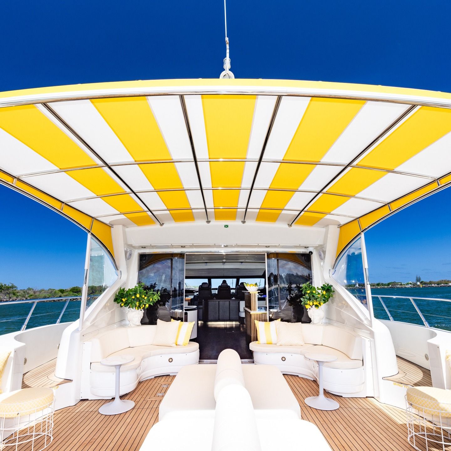 The back deck of a yacht with white cushioned seating and a yellow and white striped canopy under a clear blue sky.