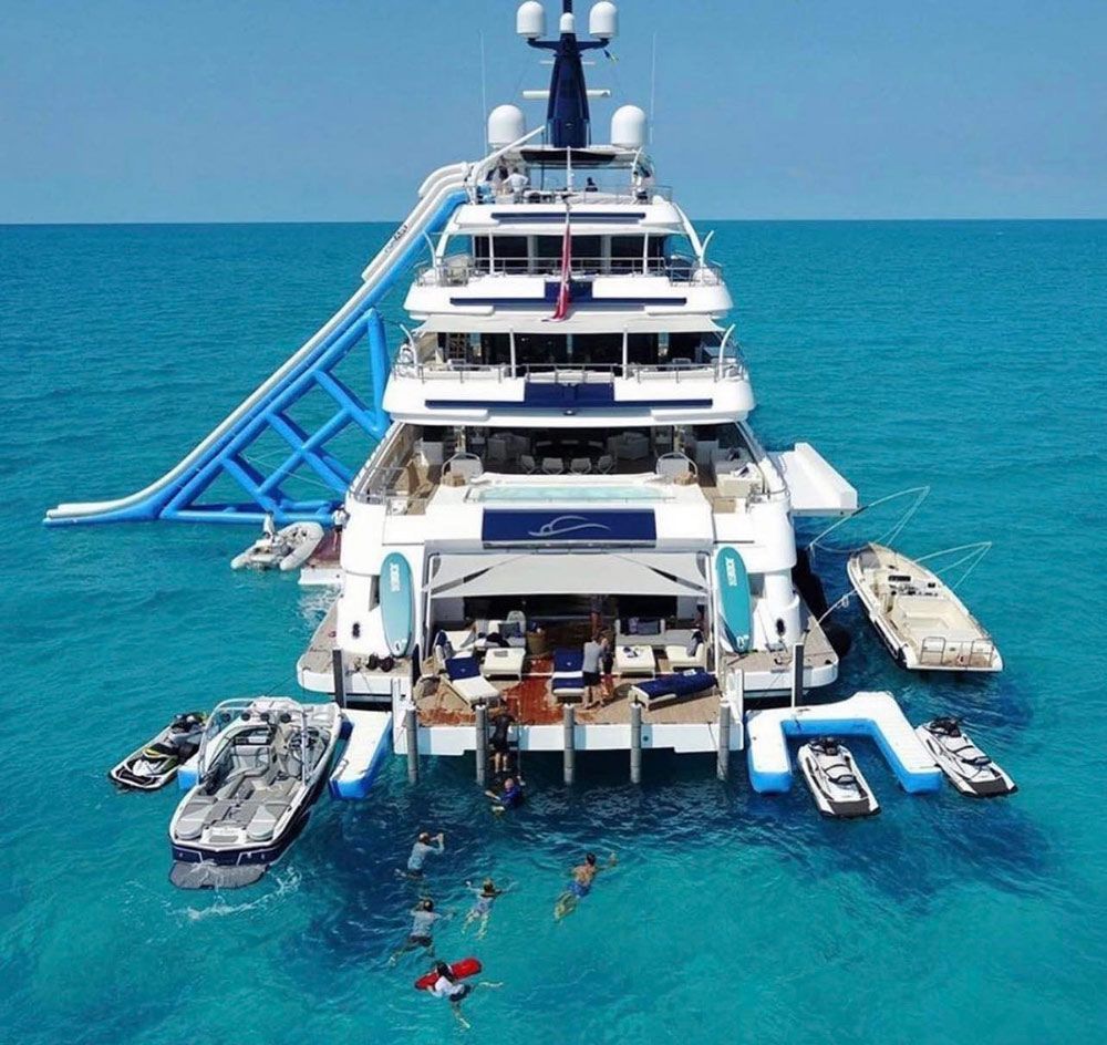 Large yacht with a blue slide, small boats, and people swimming in bright, turquoise tropical waters.
