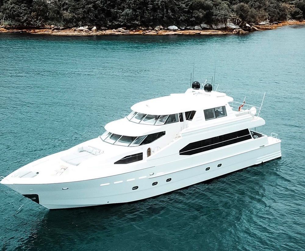A sleek white motor yacht cruises across calm, deep blue water with a wooded shoreline in the background.