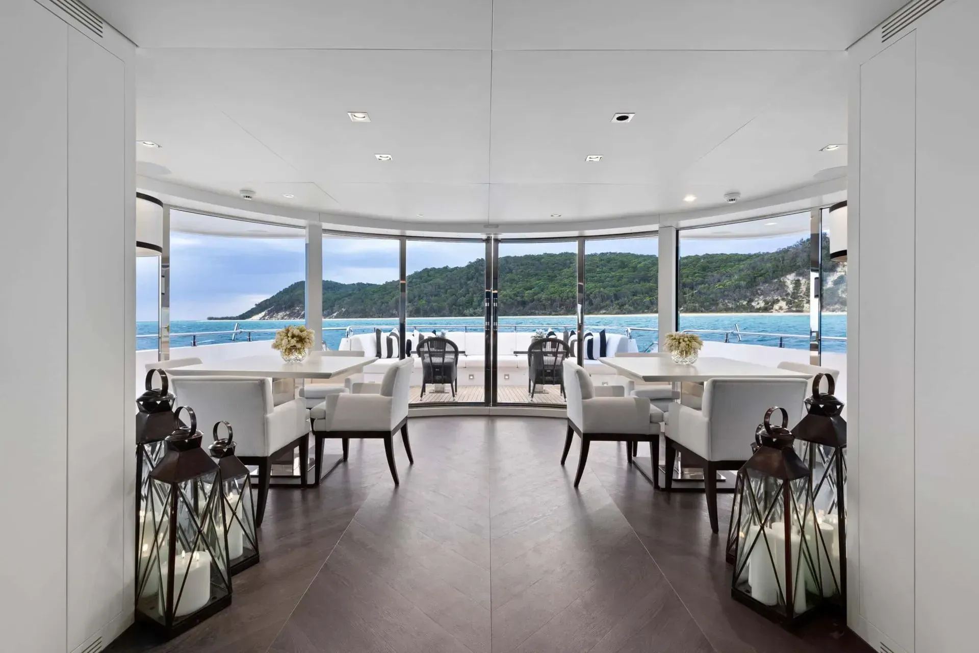 A bright, modern indoor dining area with white chairs and tables looking out through glass walls at a bay and hills.