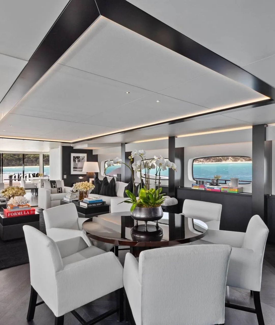 A modern yacht dining area with white chairs around a circular table, large windows, and contemporary dark trim ceilings.