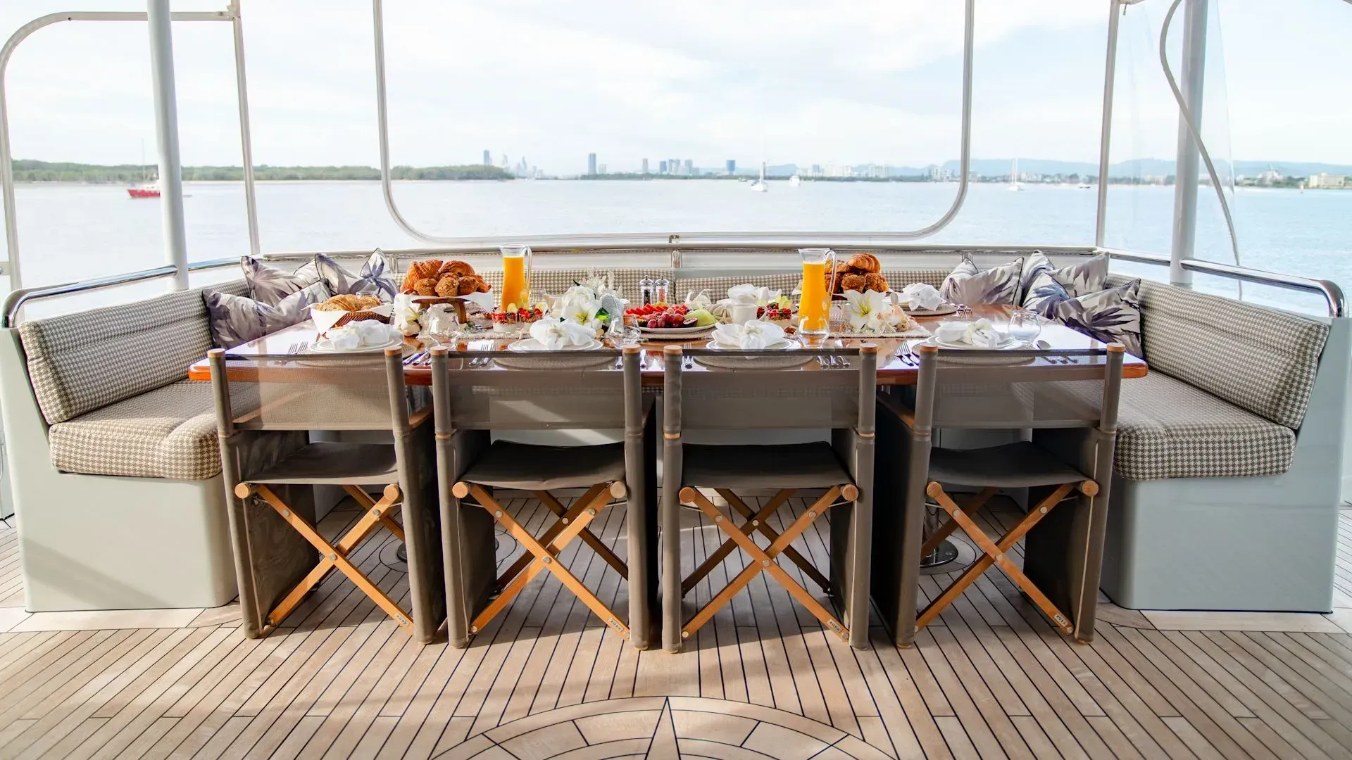An outdoor dining area on a yacht deck features a set table with food, chairs, and cushioned benches overlooking the water.