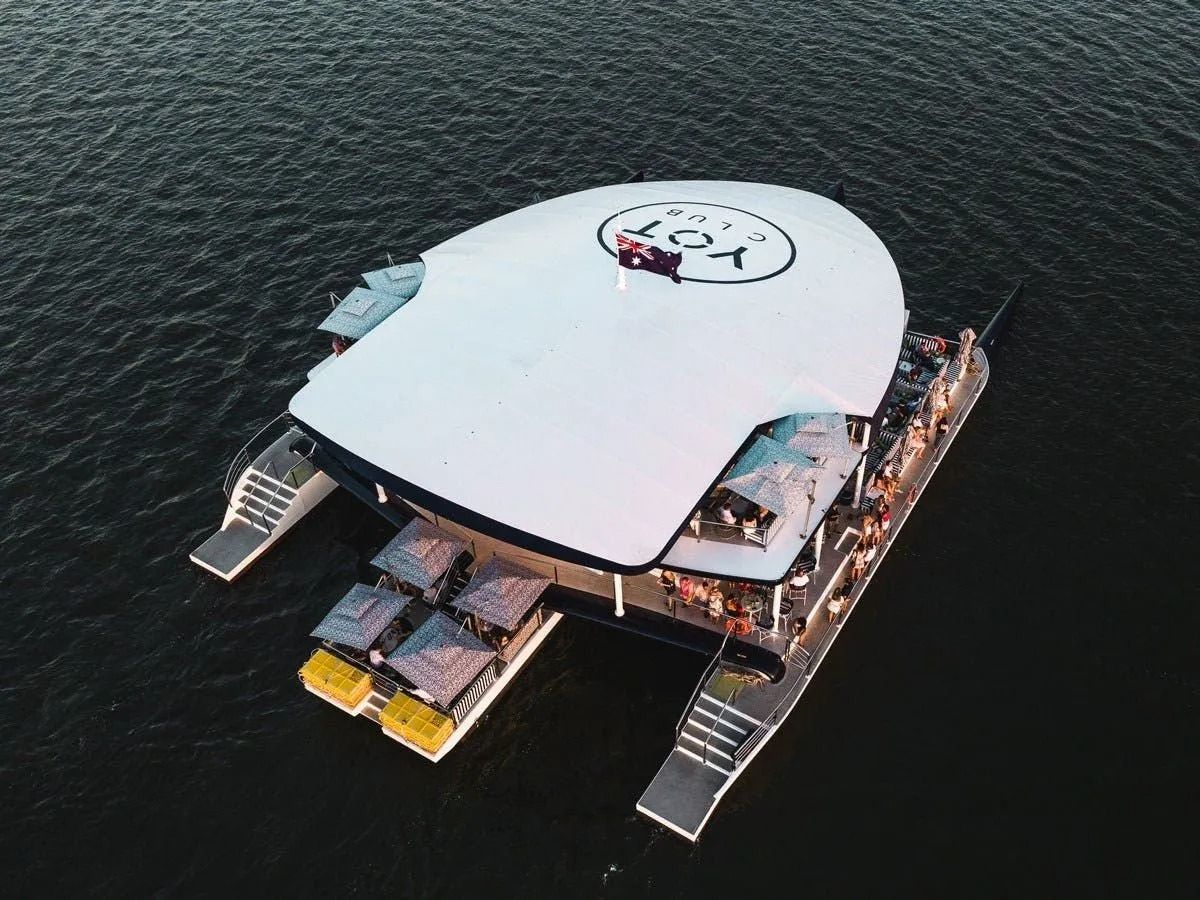 An aerial view of a large white catamaran boat on the water, featuring a branded roof and a deck filled with people.