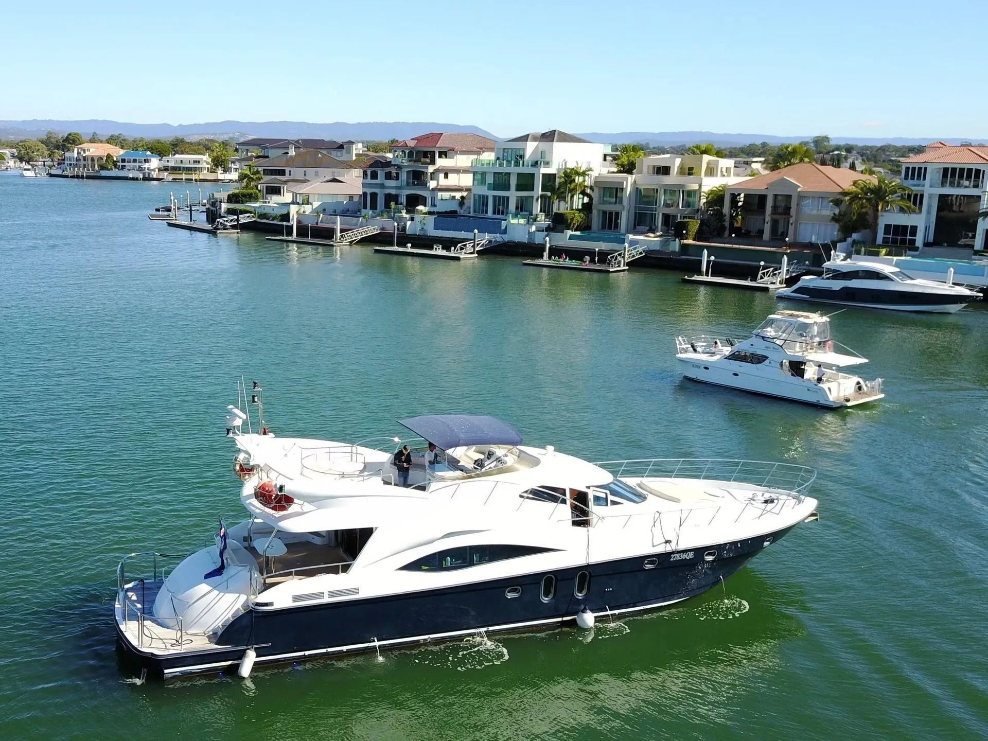 A white and navy yacht cruises on a sunny, calm waterway lined with modern houses and private docks.