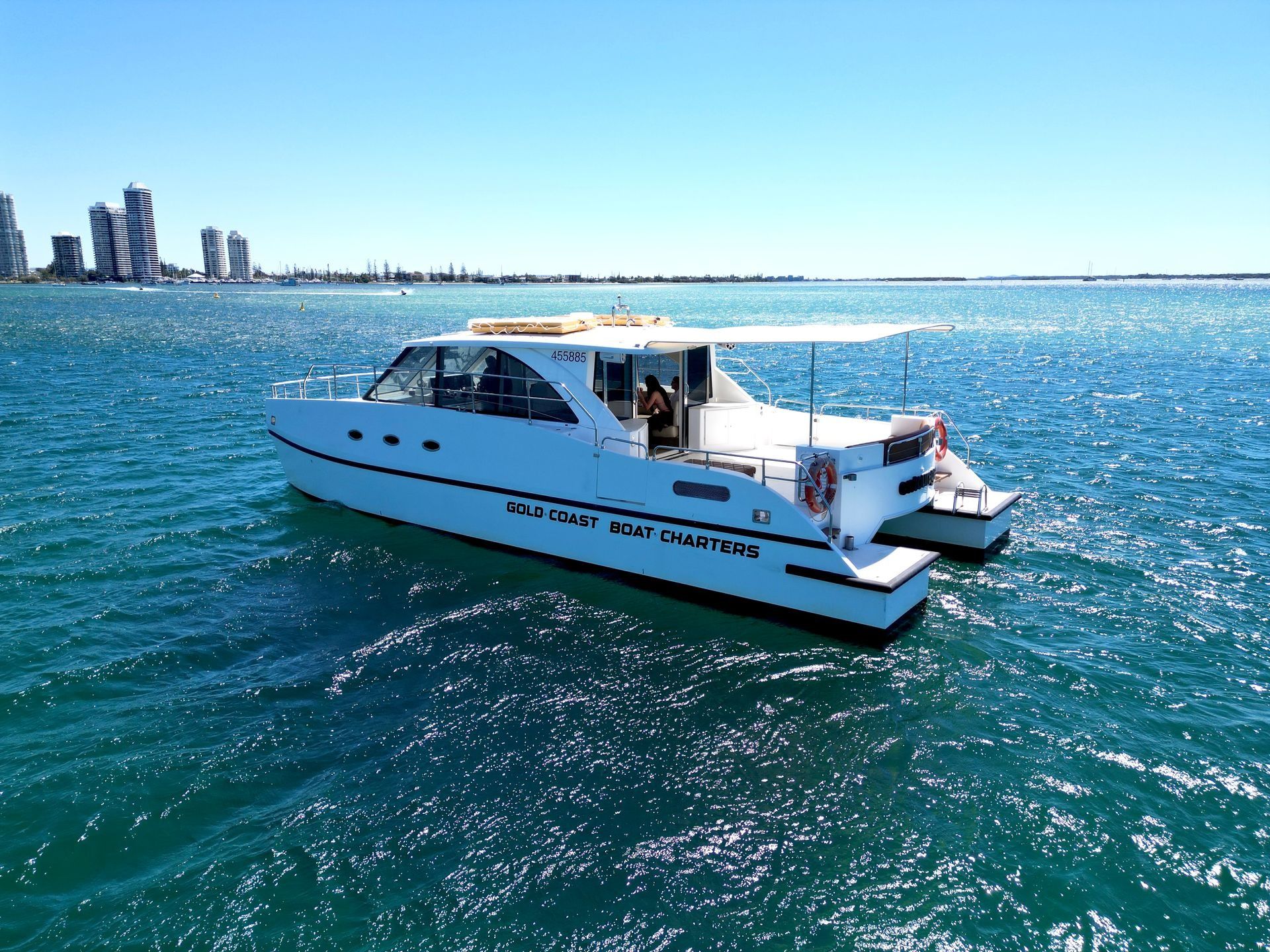 A white catamaran boat cruising on bright blue water with a city skyline visible in the distant background.