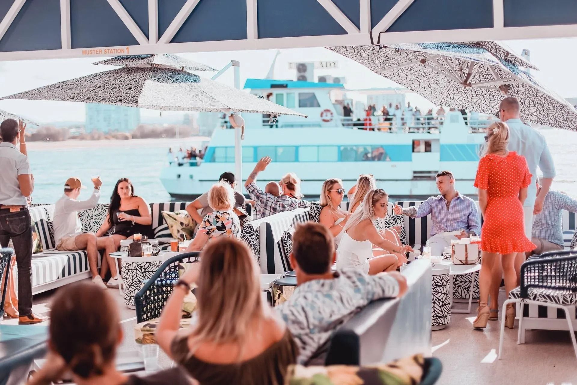 A group of people relaxes at an outdoor waterfront restaurant with a large blue boat docked in the background.