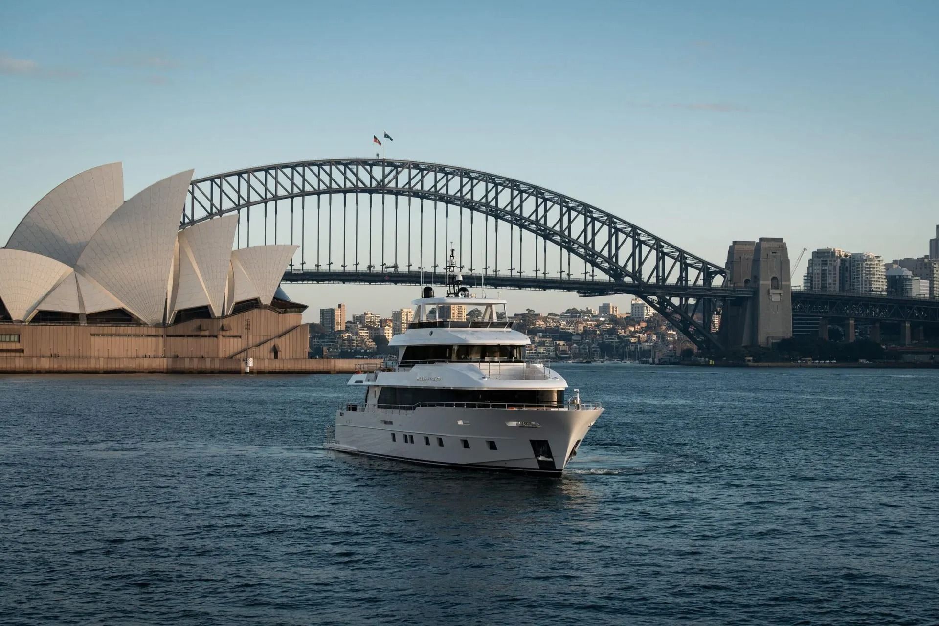 A white motor yacht cruises on the water in front of the Sydney Opera House and the Sydney Harbour Bridge.