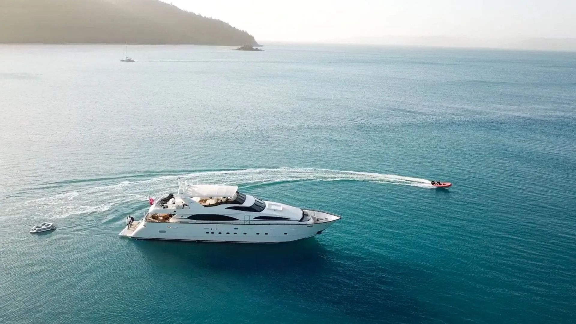 A sleek white yacht cruises through calm, deep blue water with a small boat trailing behind near a distant coastline.