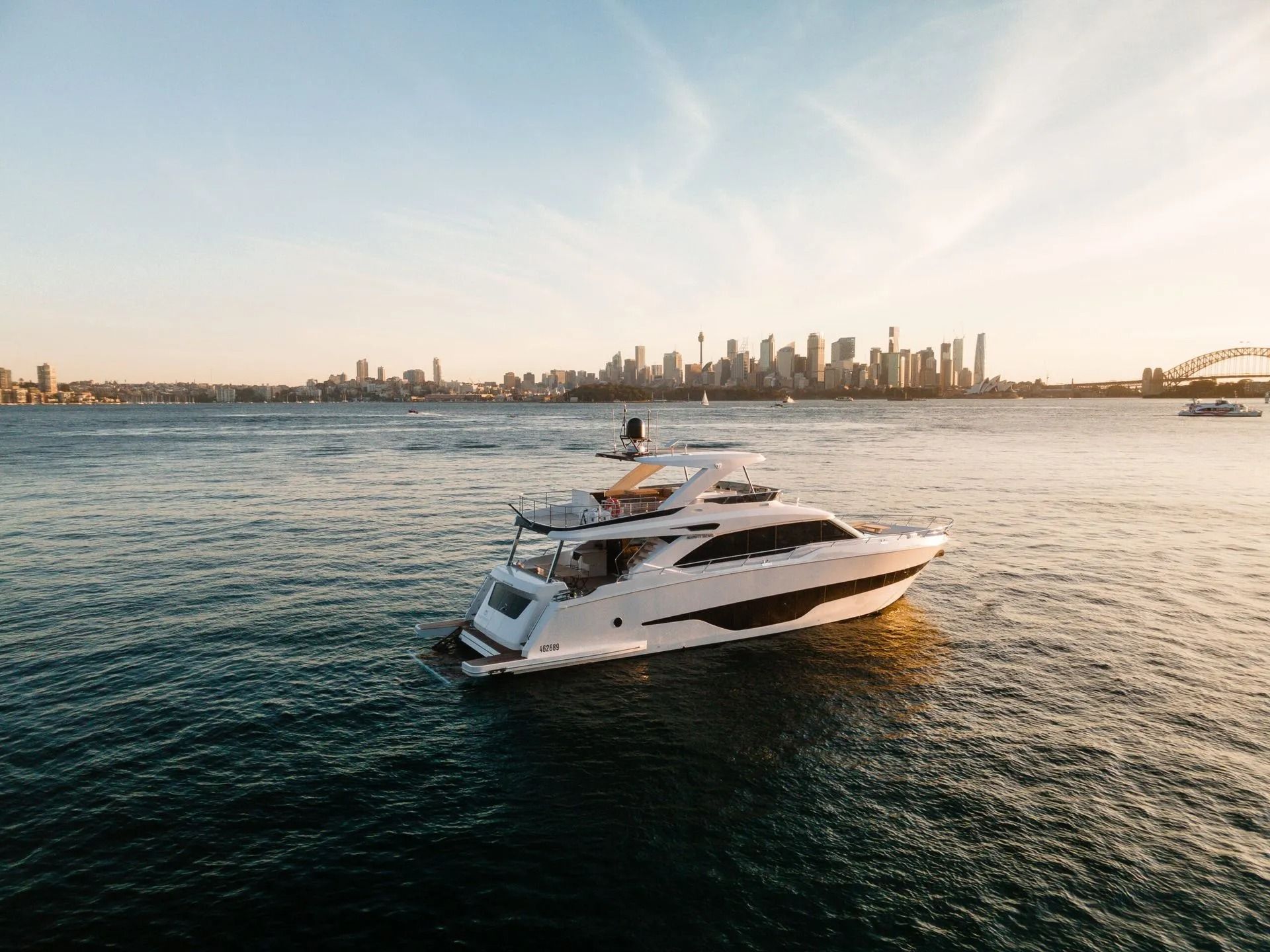 A white yacht cruises on the water with the Sydney skyline and Harbour Bridge in the background during sunset.