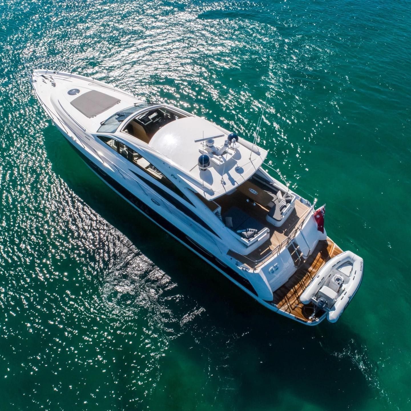 An aerial view of a sleek white yacht with a small tender boat attached, sailing on calm, clear turquoise water.