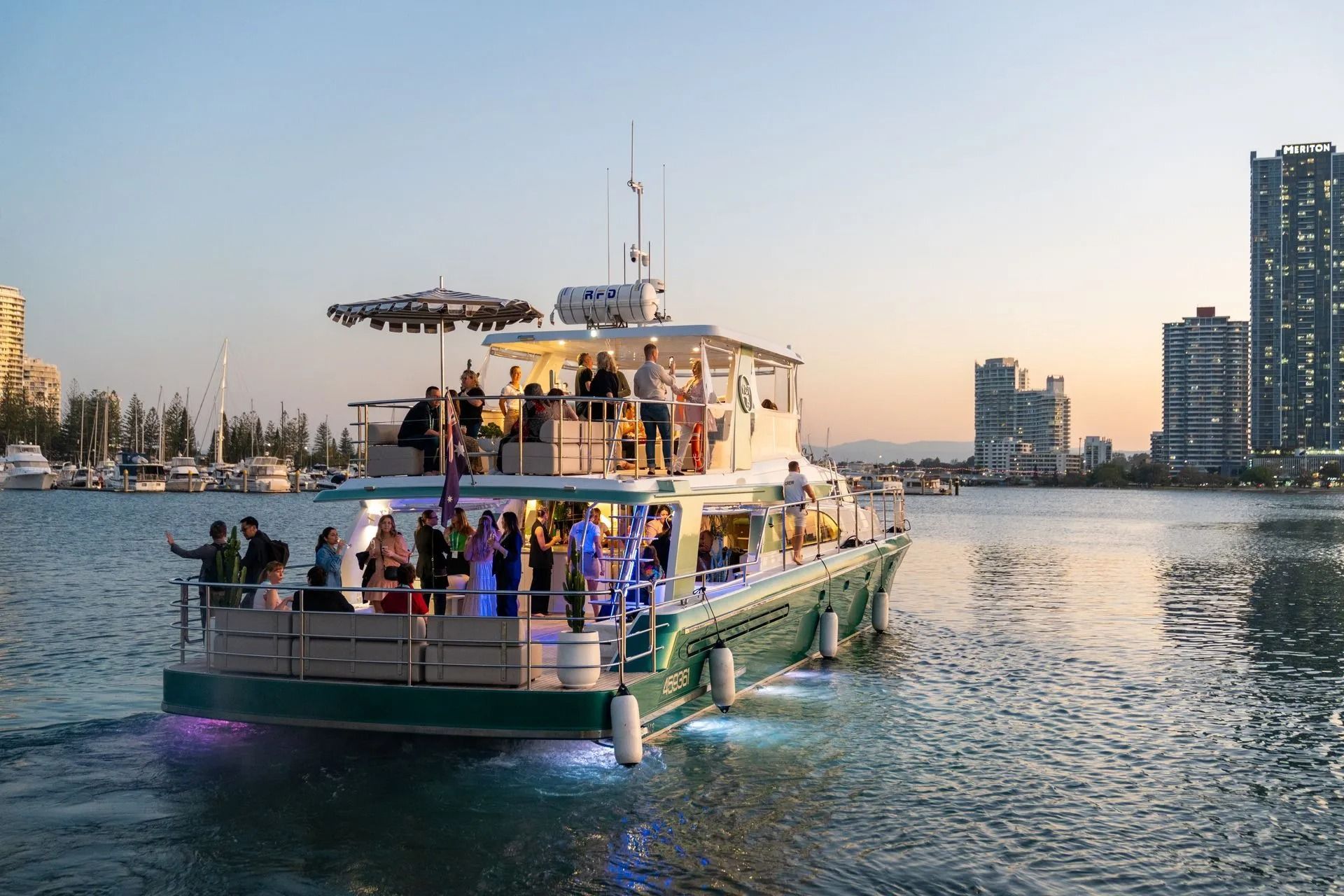 A group of people gathers for a party on a two-story boat cruising on water with a city skyline at sunset in the background.