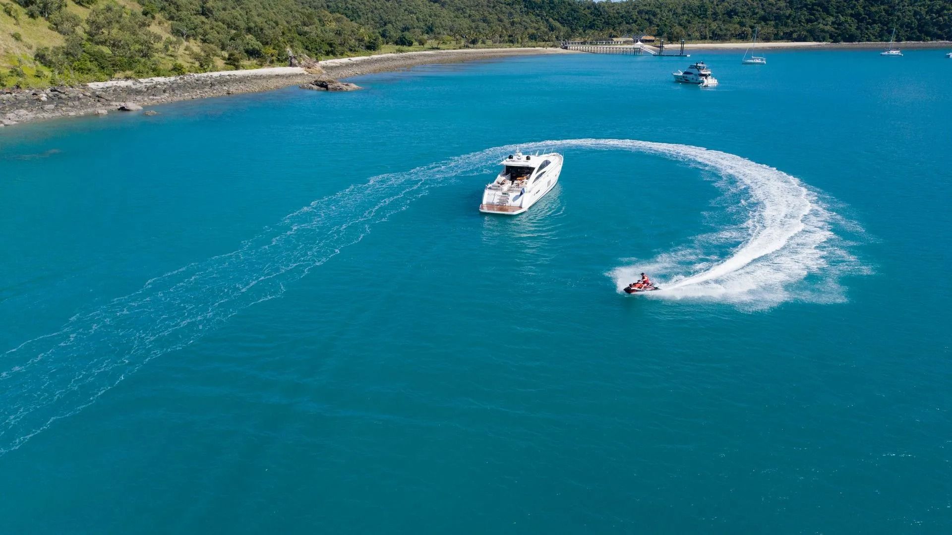 A jet ski circles a white yacht in vibrant turquoise tropical waters near a coastline.