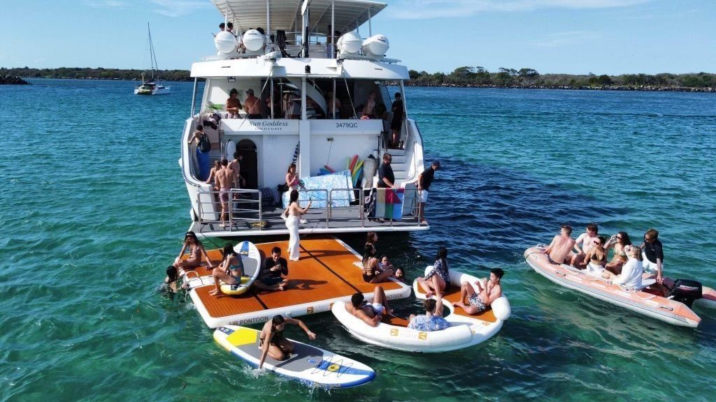 People enjoy leisure activities on a large yacht, a floating platform, a paddleboard, and an inflatable raft in the ocean.