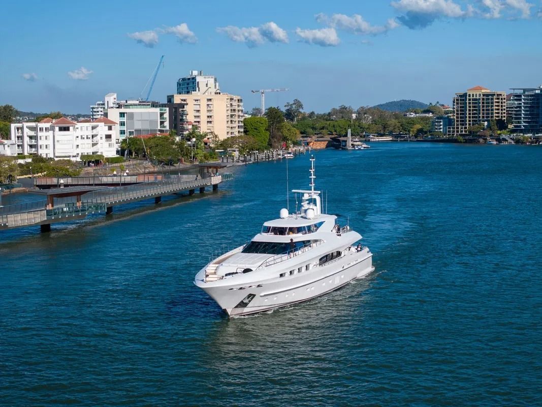 A white luxury yacht cruises through a blue waterway past a city shoreline with buildings and a wooden pier.