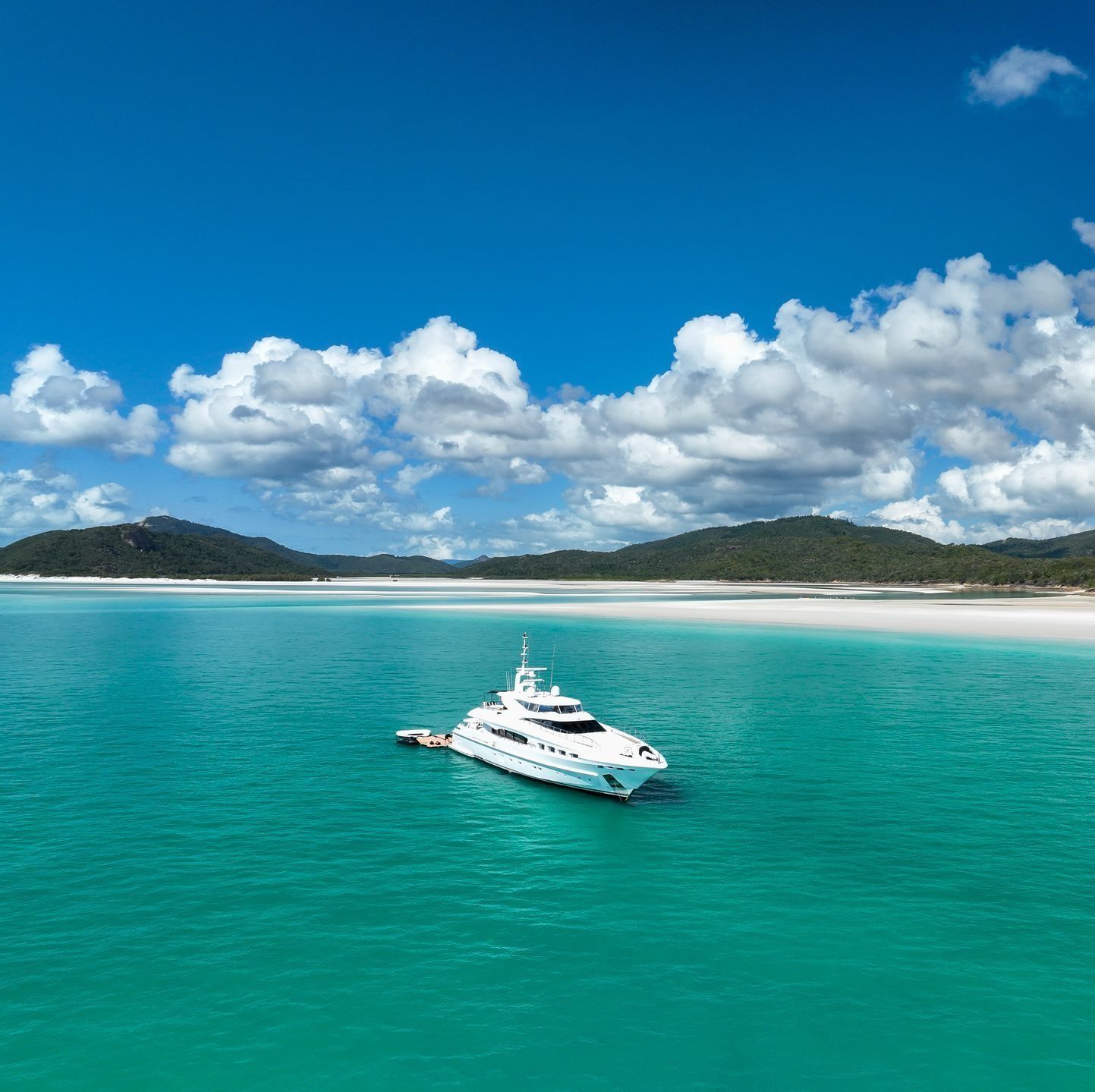 A large white yacht anchored in the bright turquoise waters of a tranquil beach with white sand and hills under blue skies.