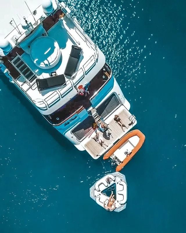 Aerial view of a white yacht on blue water with an orange dinghy and a small floating platform nearby.