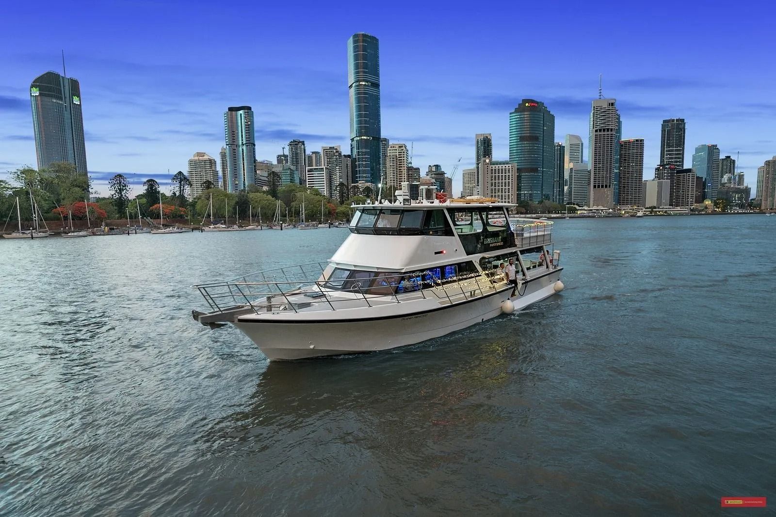 A white boat cruises on a river in front of a modern city skyline under a clear blue sky.
