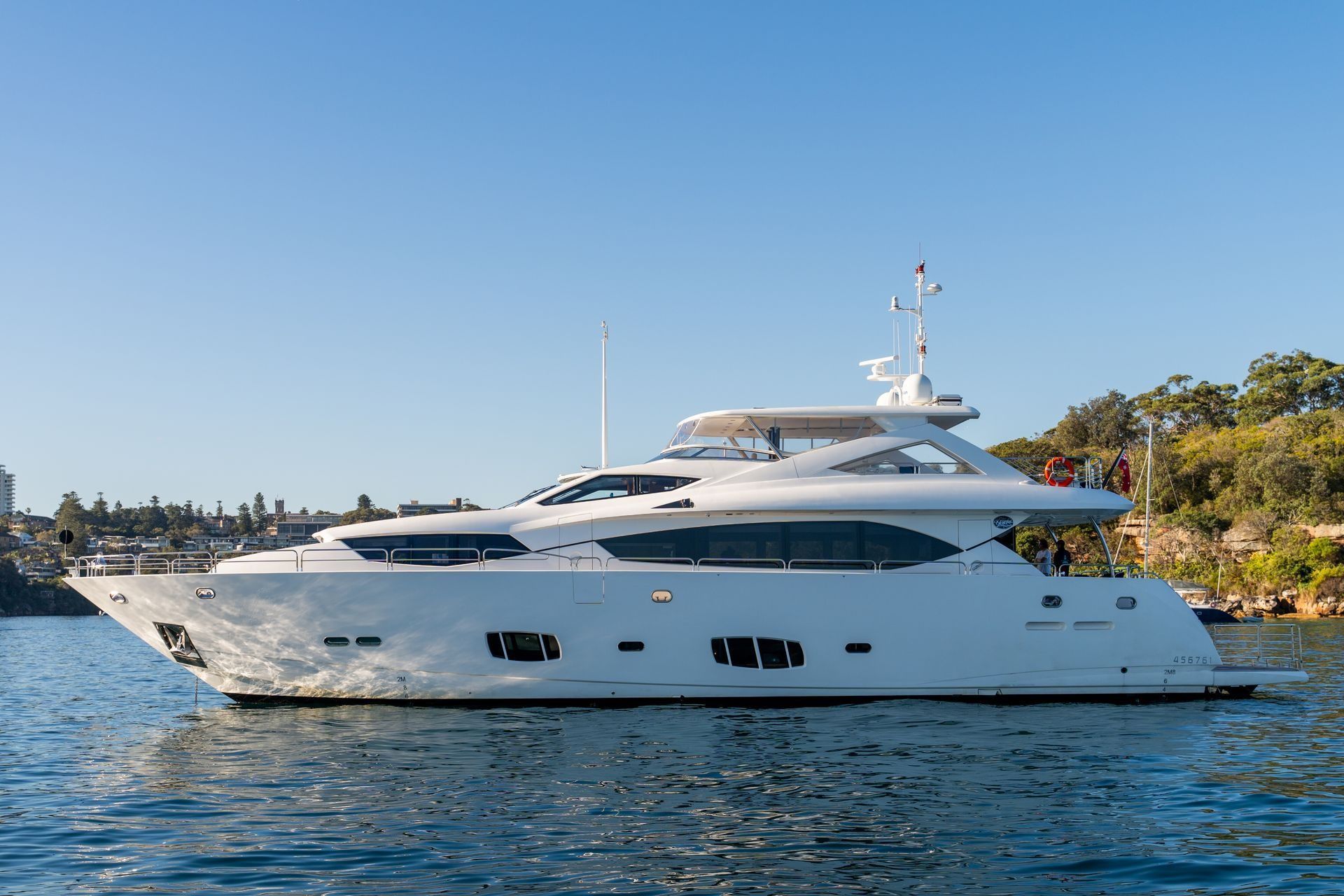 A sleek white motor yacht cruises on calm water against a clear blue sky, with a shoreline visible in the background.