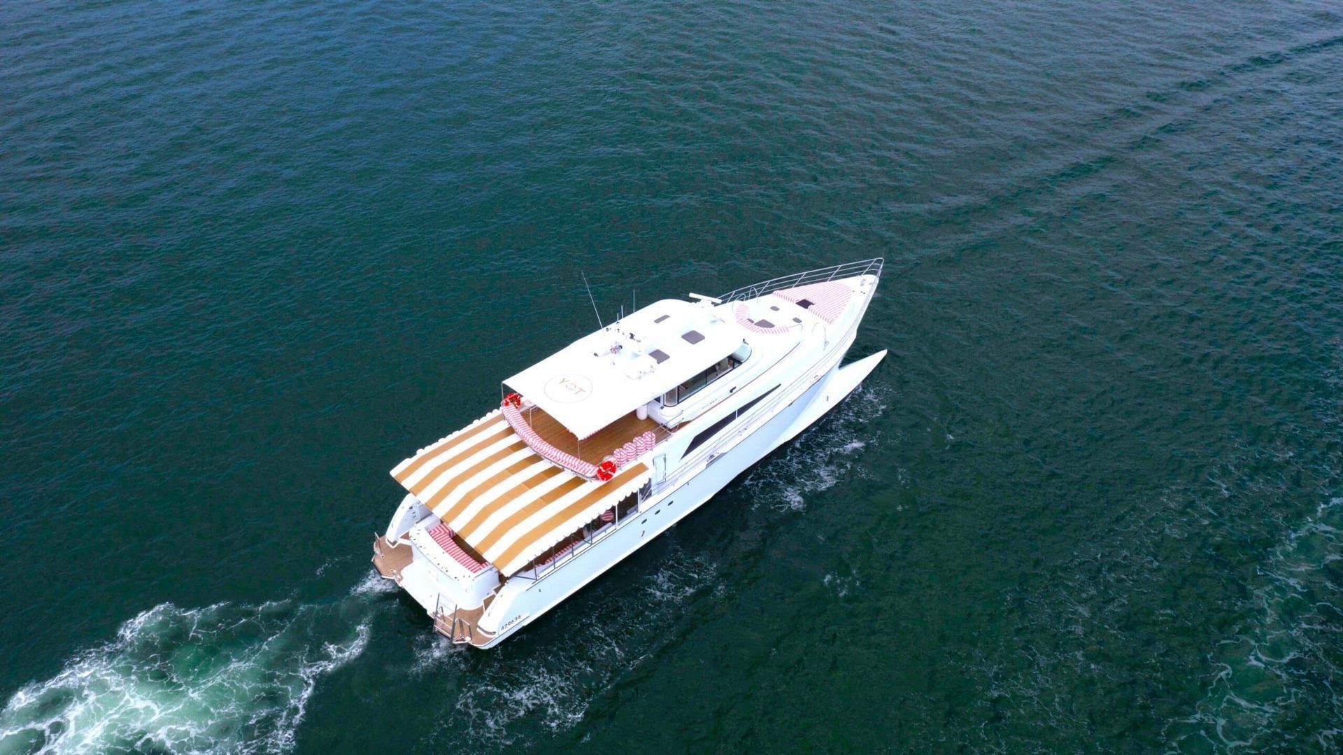 Aerial view of a white yacht with a beige-striped canopy moving through dark blue ocean water.
