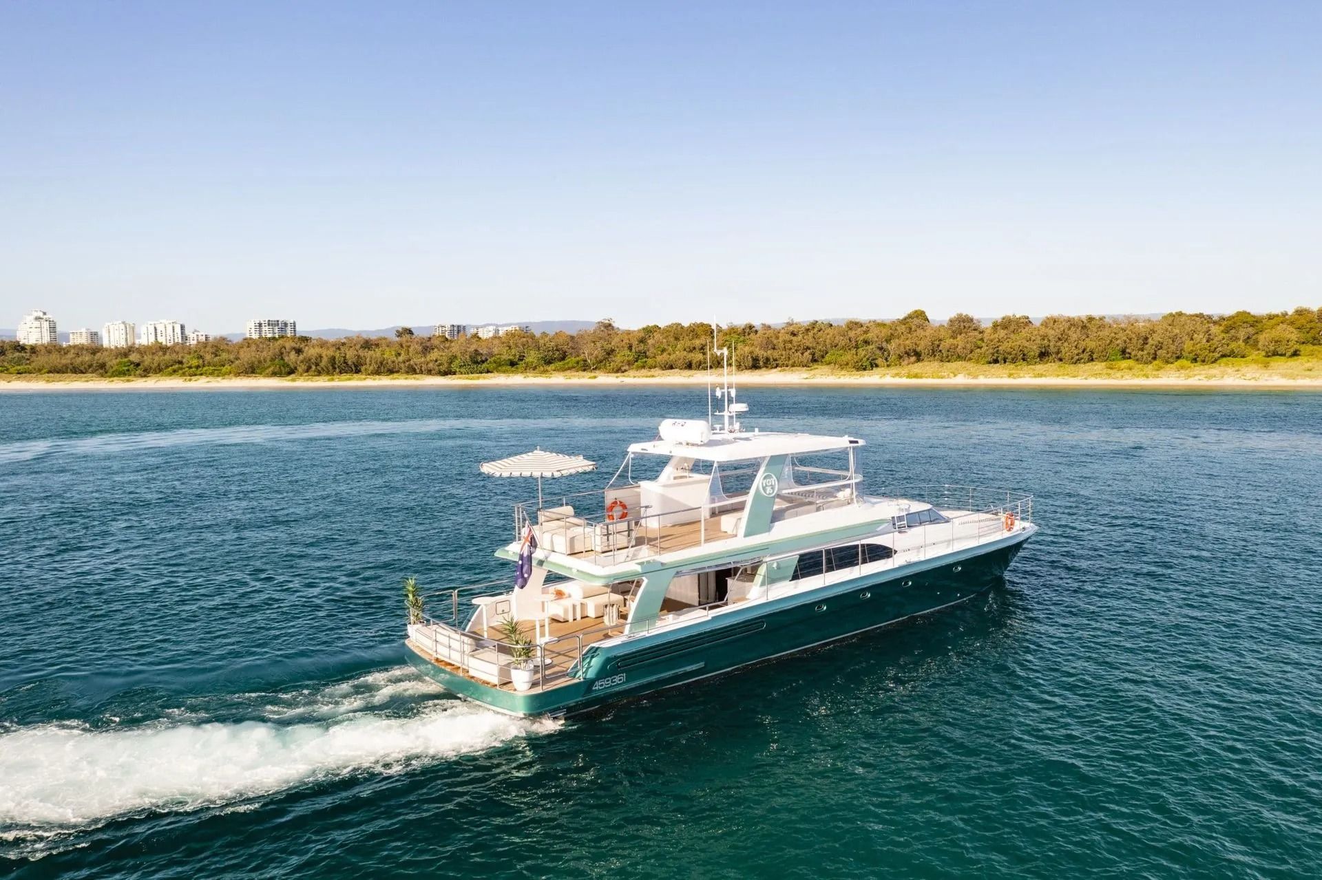 A teal and white motor yacht cruising on blue water near a tree-lined coast on a clear day.