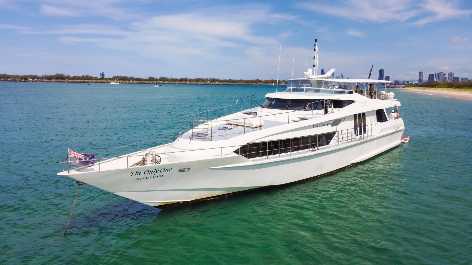 A white luxury yacht anchored in clear turquoise water near a sandy beach with a city skyline in the distance.