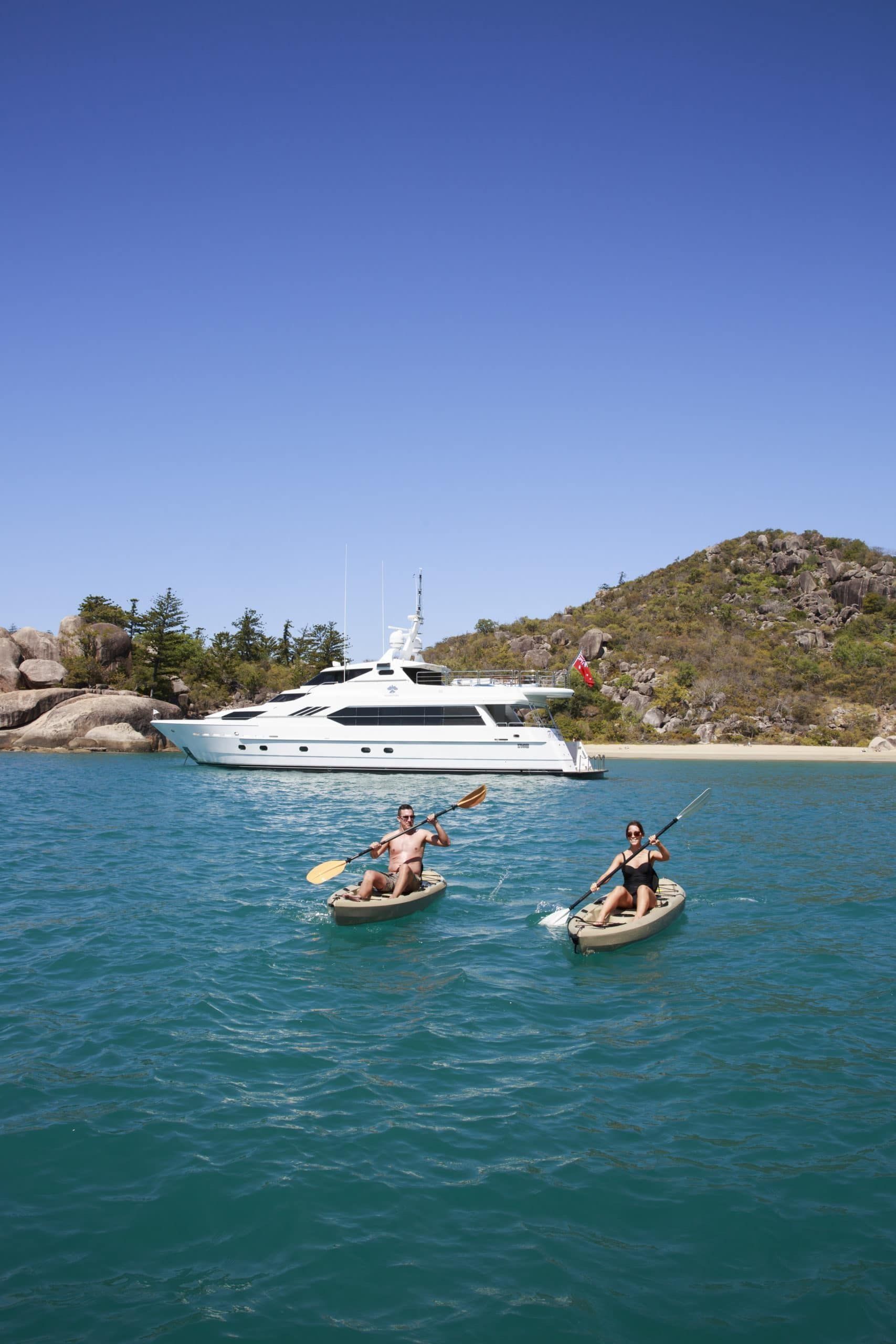 Two people kayak on turquoise water in front of a white yacht near a rocky, tree-lined coast under a clear blue sky.
