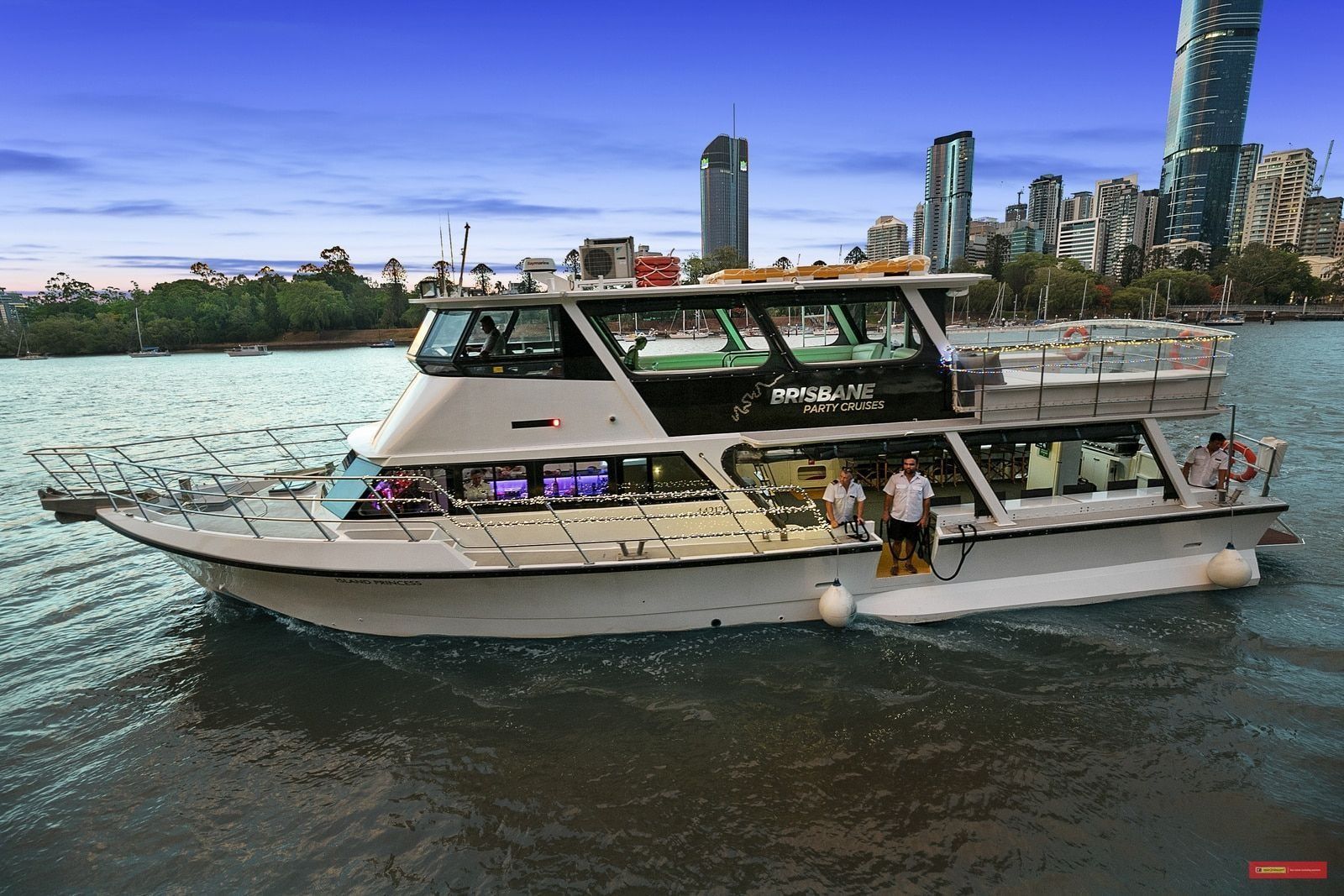 A white multi-level river cruise boat sailing on a waterway with a city skyline in the background.
