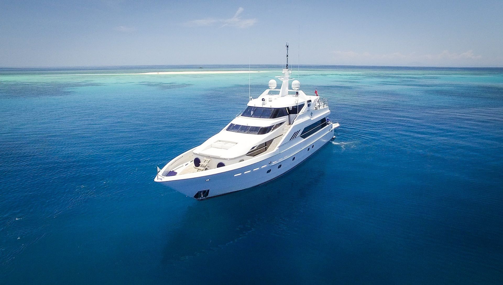 A sleek white yacht sits anchored in the middle of bright blue tropical waters near a shallow reef.
