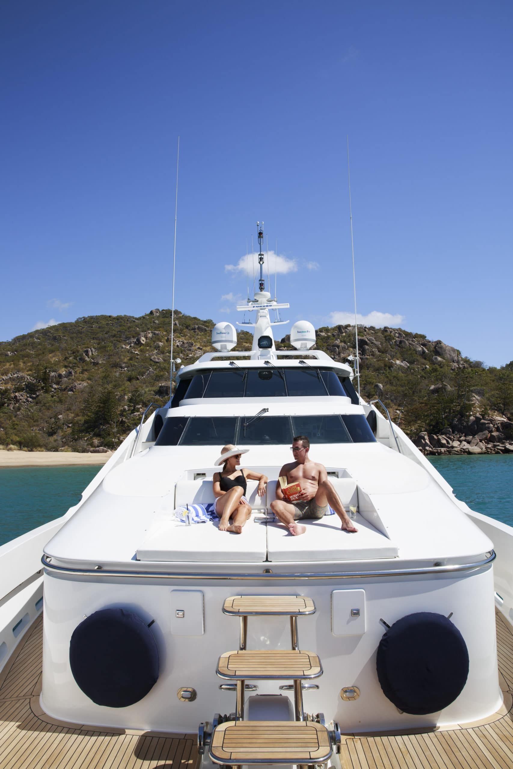 Two people sunbathing on white lounge cushions on the bow of a yacht docked in a tropical bay.