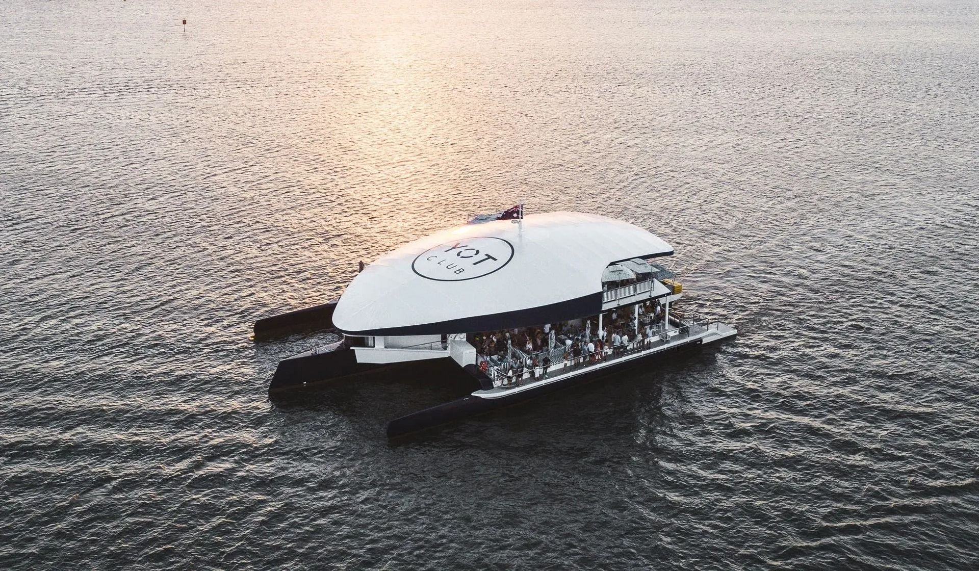 An aerial view of a white, oval-shaped floating venue or boat on open water, with people gathered on the rear deck.