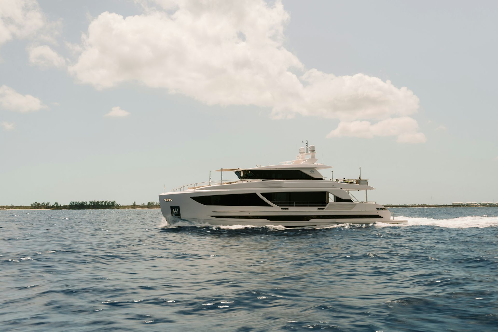 A sleek white motor yacht cruises across open blue water under a partly cloudy sky.