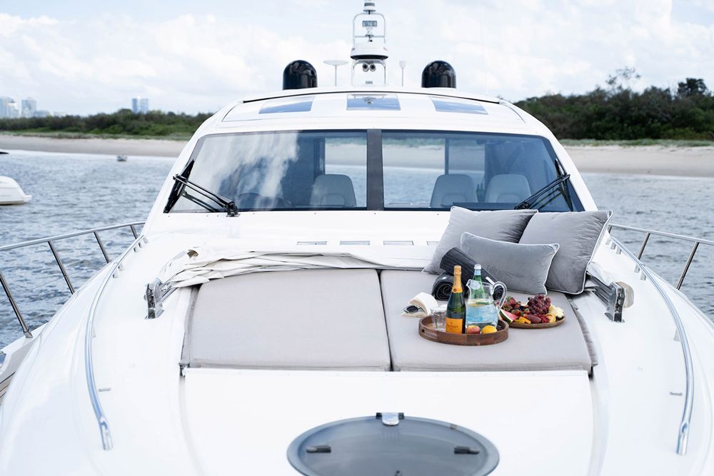 A white yacht’s sun deck on the water, featuring grey cushions, pillows, a bottle of champagne, and a fruit platter.