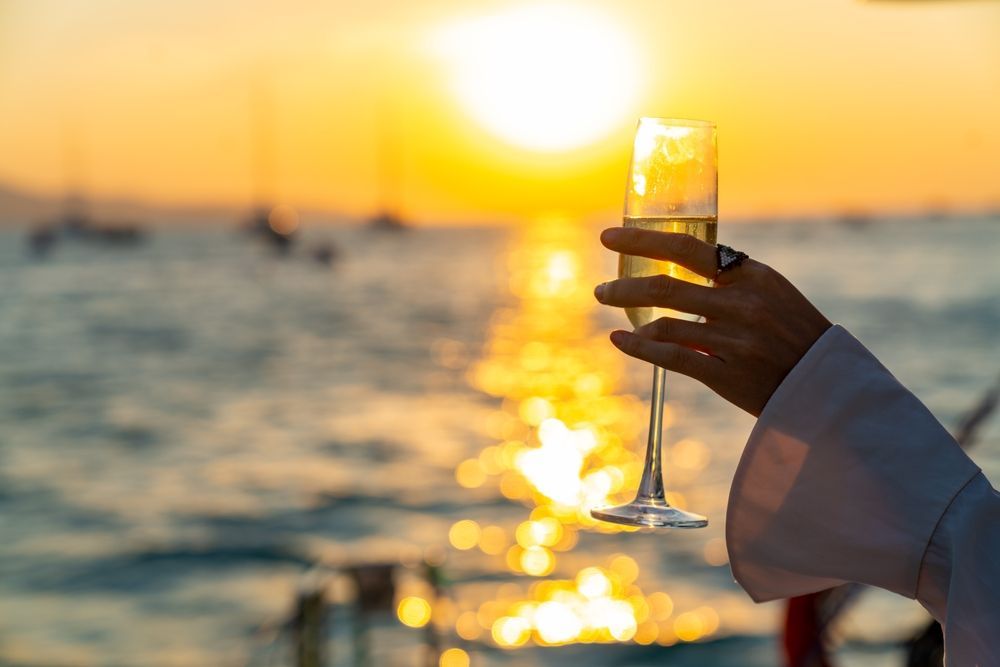 A hand holds a glass of champagne against a bright golden sunset over the ocean, with silhouettes of sailboats.