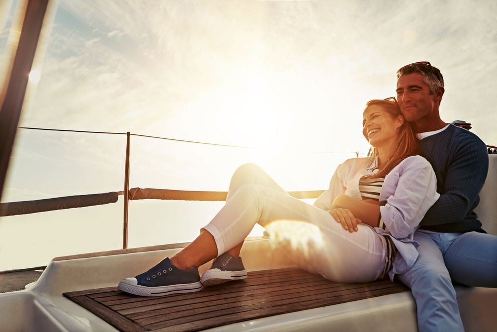 A couple sits closely together on the deck of a sailboat at sunset, looking out toward the horizon.