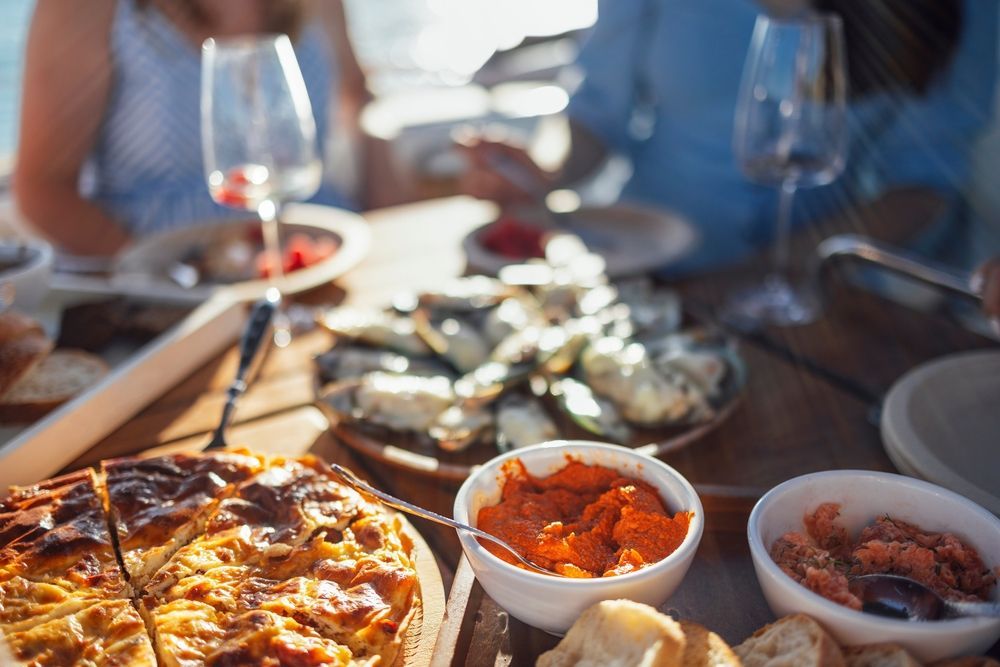 People dining outdoors on a wooden table with a quiche, bowls of spread, and a platter of oysters.