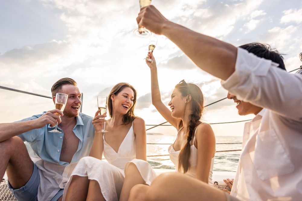 Four people sitting on a boat deck during sunset, laughing and raising their glasses in a toast.