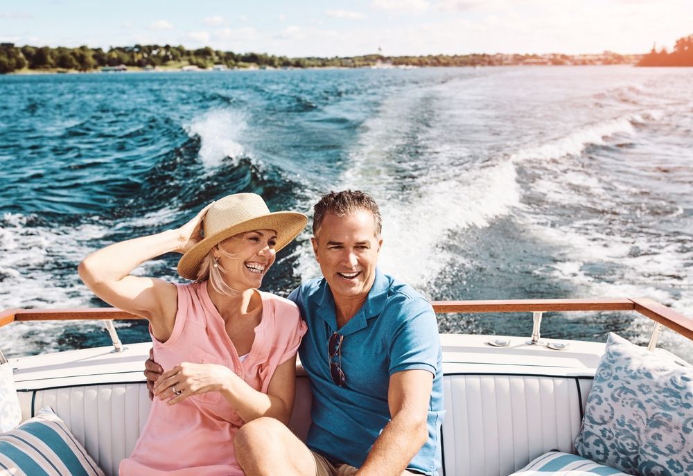 A smiling couple sits on a boat with a wake trailing behind them on a sunny day.