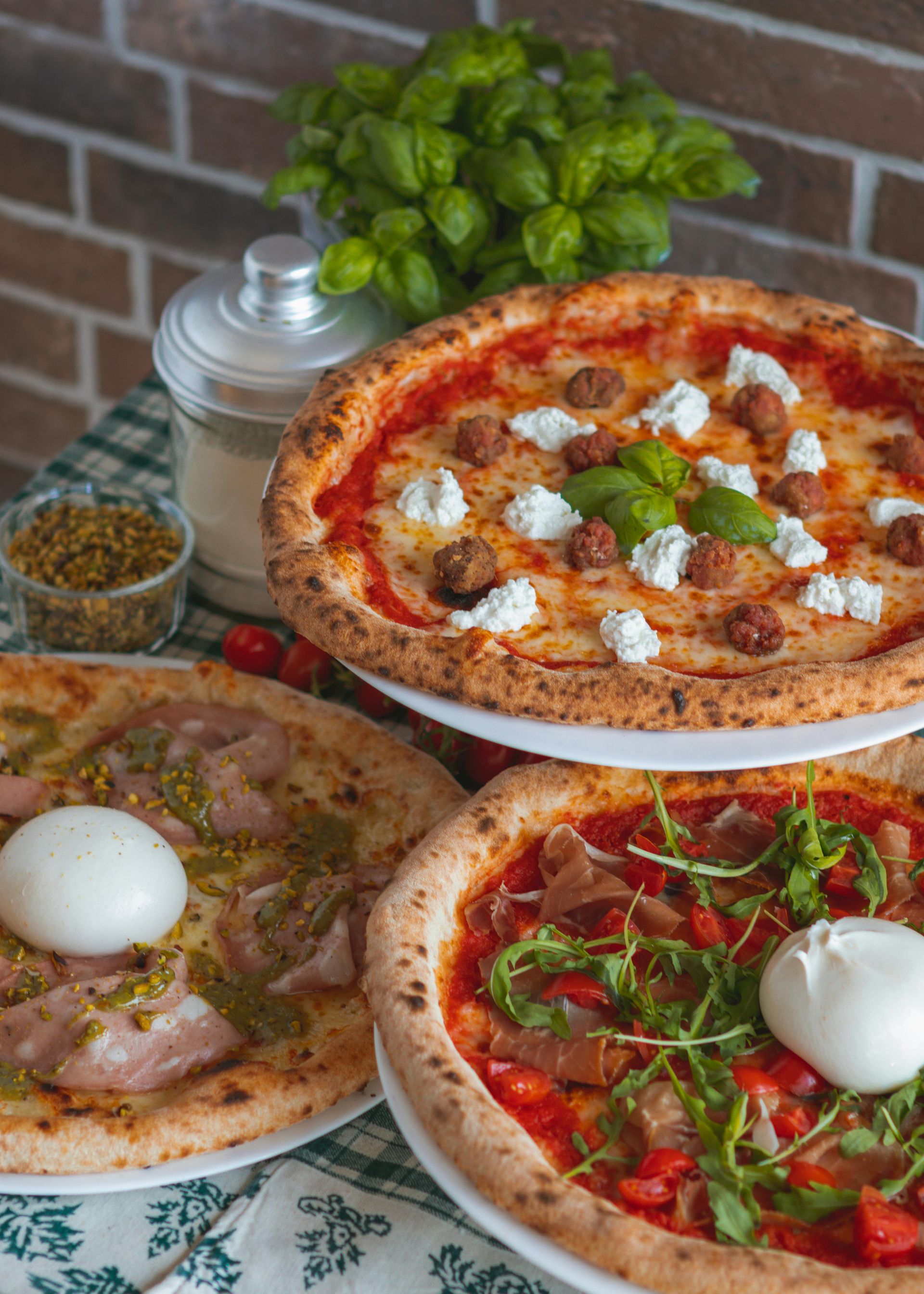 Three pizzas with various toppings, including meatballs, ricotta, and prosciutto, on a table with basil and spices.